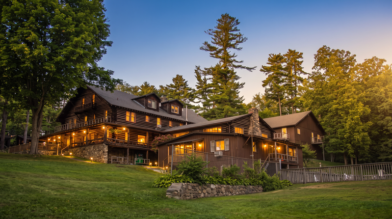 A large log cabin-style building with lights on, surrounded by trees and a grassy yard.