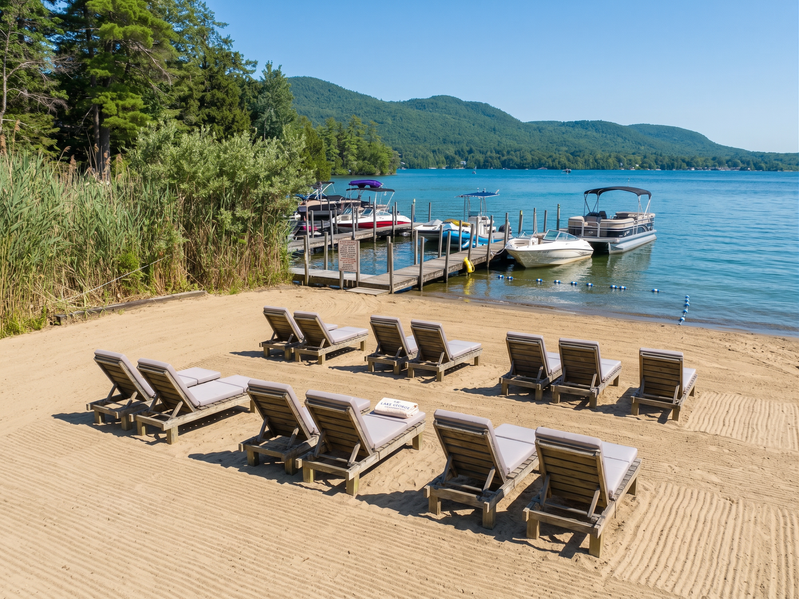 A sandy beach with lounge chairs, a dock, and boats on a lake with mountains in the background.