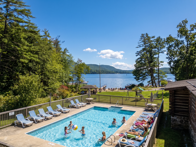 A rectangular swimming pool with people in it, surrounded by lounge chairs and trees, with a lake in the background.